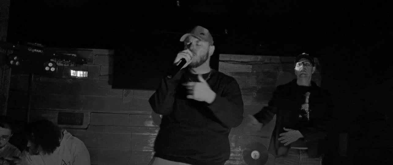 Person speaking into a microphone with another person standing nearby in a dimly lit room.